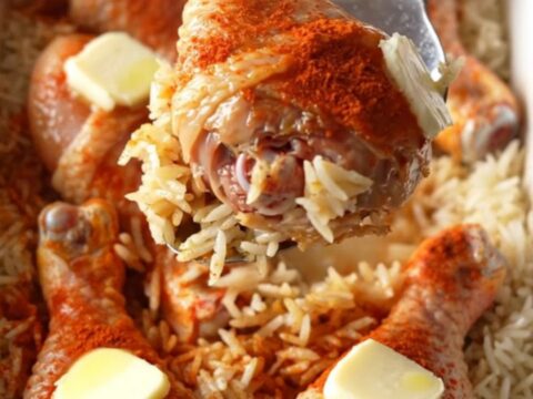 A top-down view of raw chicken drumsticks rubbed with red paprika sitting on a bed of white rice in a white baking dish.