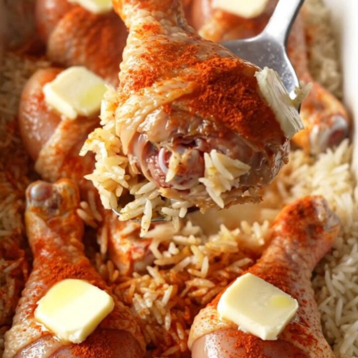 A top-down view of raw chicken drumsticks rubbed with red paprika sitting on a bed of white rice in a white baking dish.