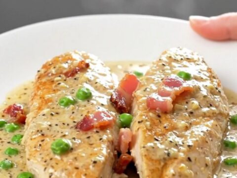 A close-up of tender, braised chicken breasts in a creamy, peppery Parmesan sauce with bacon and peas.