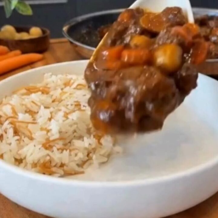 A wide white bowl filled with glossy slow-cooked beef stew, tender carrots, and potatoes over fluffy vermicelli rice.