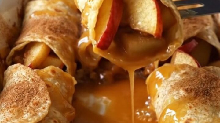 A top-down view of cinnamon apple pie enchiladas in a white baking dish, showing golden brown rolls and bubbling syrup.