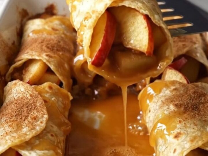 A top-down view of cinnamon apple pie enchiladas in a white baking dish, showing golden brown rolls and bubbling syrup.