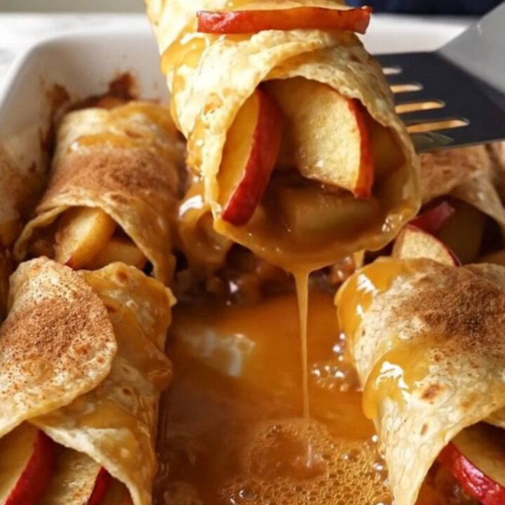 A top-down view of cinnamon apple pie enchiladas in a white baking dish, showing golden brown rolls and bubbling syrup.