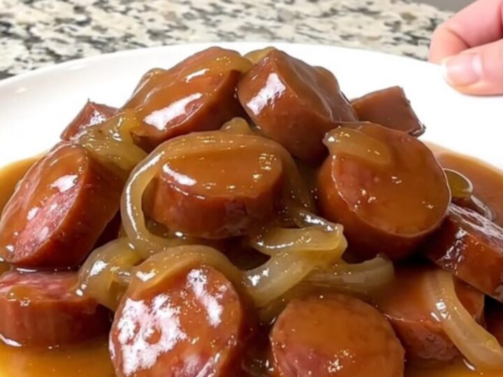 A close-up of a slow cooker filled with sliced smoked sausage, cubed gold potatoes, and onions in a rich