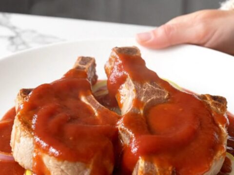 Four bone-in pork chops arranged in a slow cooker topped with sliced onions, lemons, brown sugar, and a ketchup-based sauce.