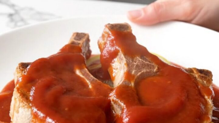 Four bone-in pork chops arranged in a slow cooker topped with sliced onions, lemons, brown sugar, and a ketchup-based sauce.