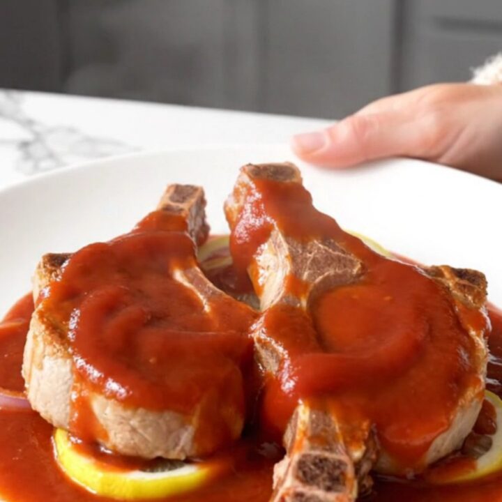 Four bone-in pork chops arranged in a slow cooker topped with sliced onions, lemons, brown sugar, and a ketchup-based sauce.