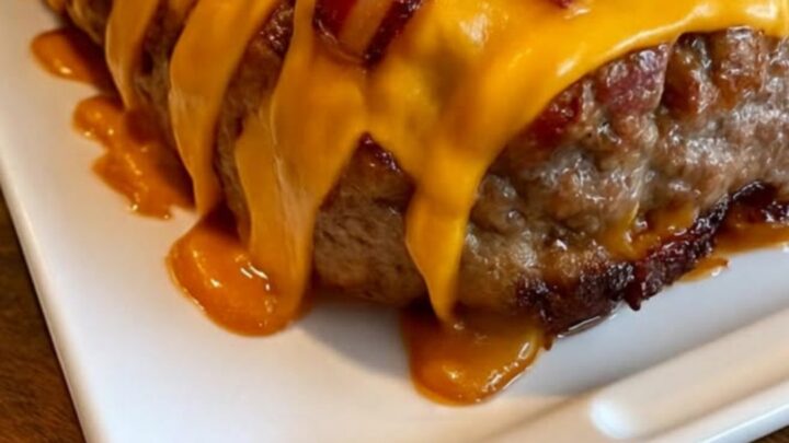 A slice of juicy meatloaf on a white platter, revealing a molten cheddar cheese center and topped with crispy bacon.