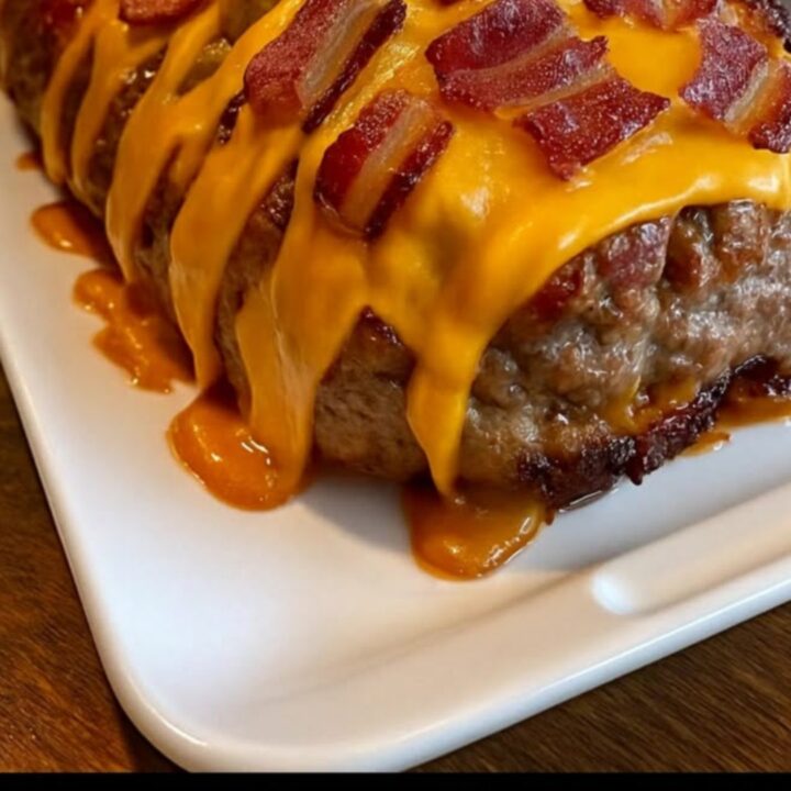 A slice of juicy meatloaf on a white platter, revealing a molten cheddar cheese center and topped with crispy bacon.
