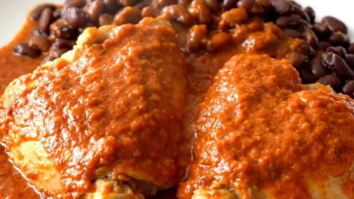 A close-up of tender chicken thighs covered in red enchilada sauce with black beans in a slow cooker.