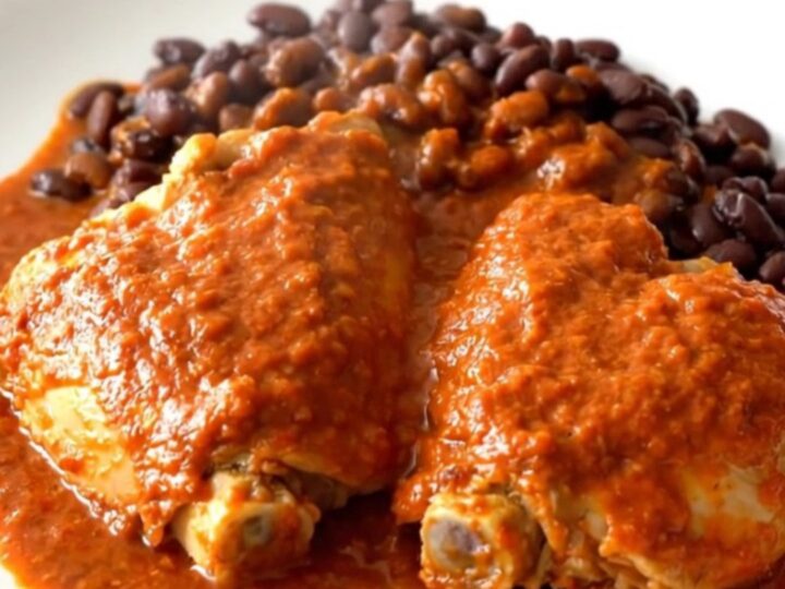 A close-up of tender chicken thighs covered in red enchilada sauce with black beans in a slow cooker.