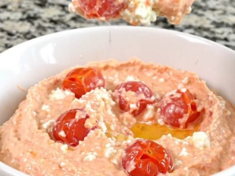 A block of feta cheese surrounded by cherry tomatoes in a black slow cooker insert on a granite countertop.
