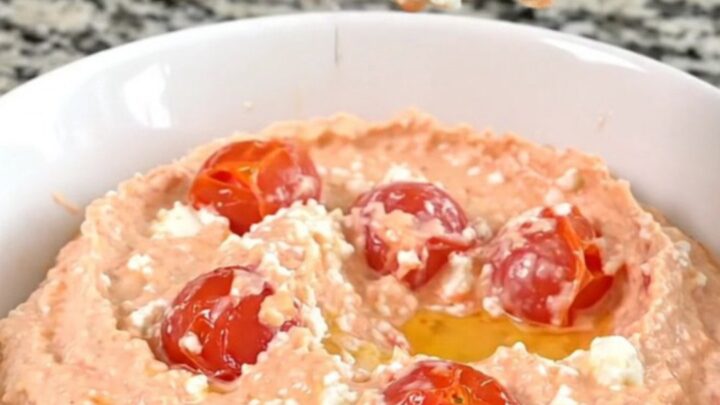 A block of feta cheese surrounded by cherry tomatoes in a black slow cooker insert on a granite countertop.