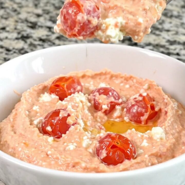 A block of feta cheese surrounded by cherry tomatoes in a black slow cooker insert on a granite countertop.