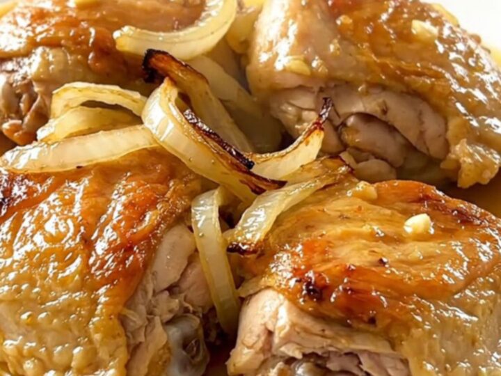 A close-up of tender, braised mojo chicken thighs topped with softened onions and citrus sauce on a white plate.