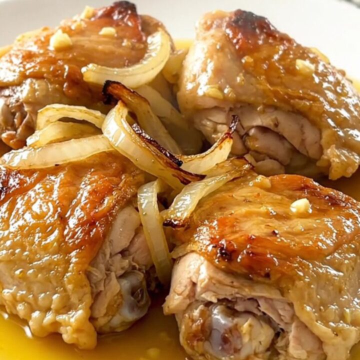 A close-up of tender, braised mojo chicken thighs topped with softened onions and citrus sauce on a white plate.