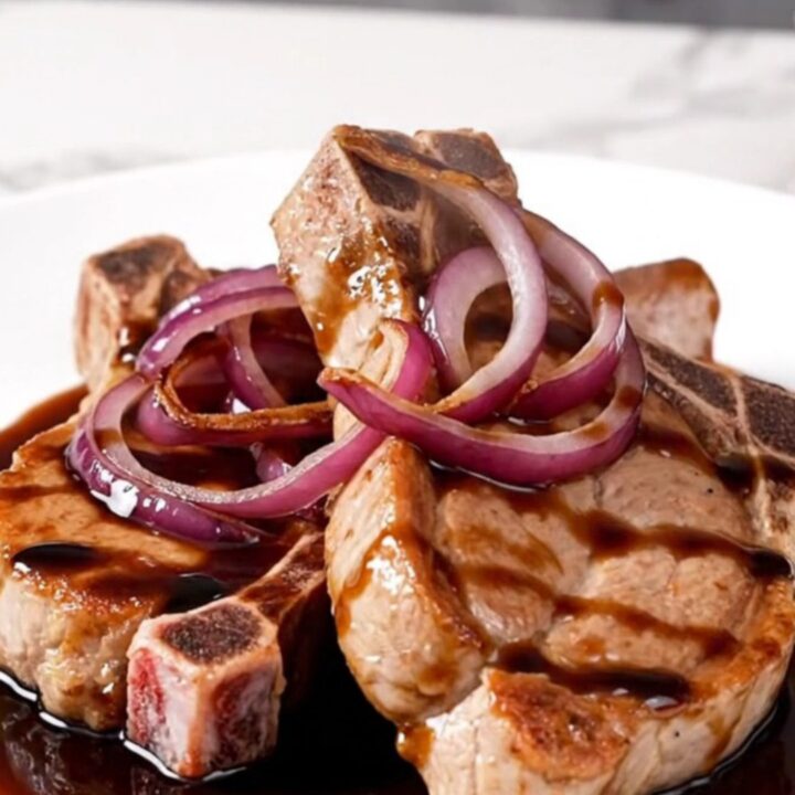 A close-up of tender, braised pork chops topped with caramelized red onions and a rich, dark balsamic sauce.