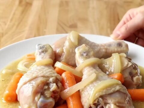 A white slow cooker filled with chicken drumsticks, carrots, and onions in a creamy sauce.