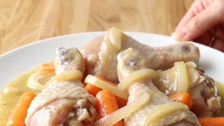 A white slow cooker filled with chicken drumsticks, carrots, and onions in a creamy sauce.