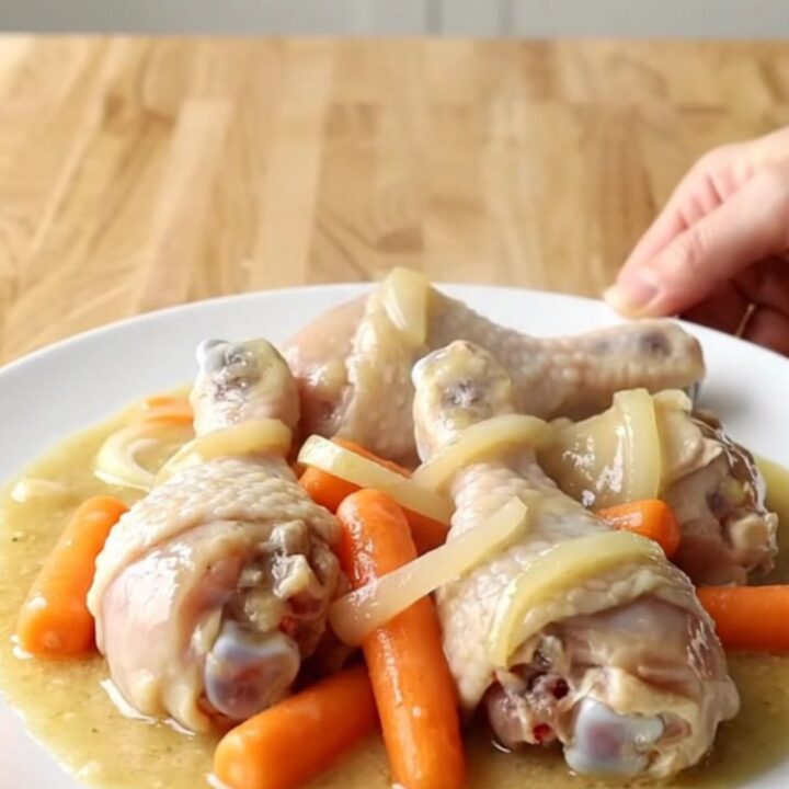 A white slow cooker filled with chicken drumsticks, carrots, and onions in a creamy sauce.