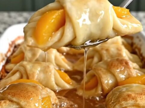 A ceramic baking dish filled with golden-brown peach crescent dumplings covered in a rich, bubbling cinnamon syrup.