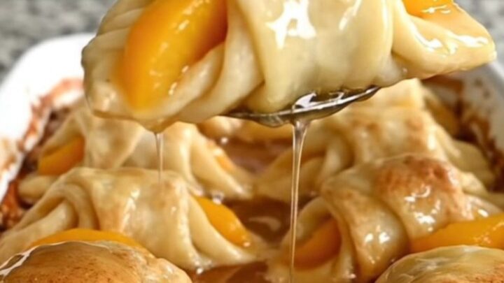 A ceramic baking dish filled with golden-brown peach crescent dumplings covered in a rich, bubbling cinnamon syrup.