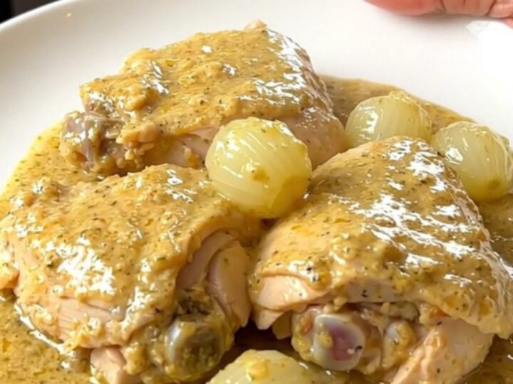 A slow cooker filled with cooked chicken thighs, whole pearl onions, and a rich, creamy white sauce with thyme.