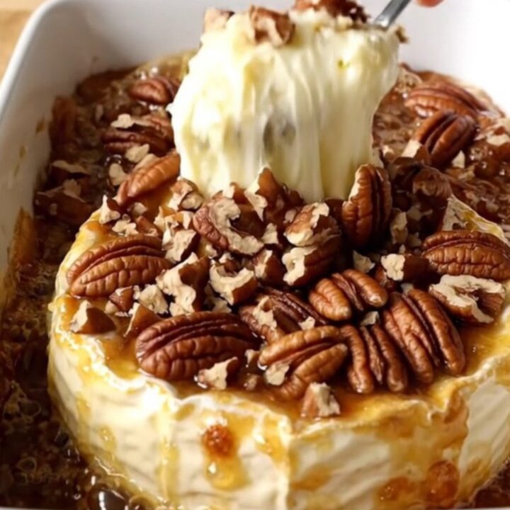 A warm, golden wheel of baked Brie in a white ceramic dish topped with caramelized brown sugar and toasted pecans.