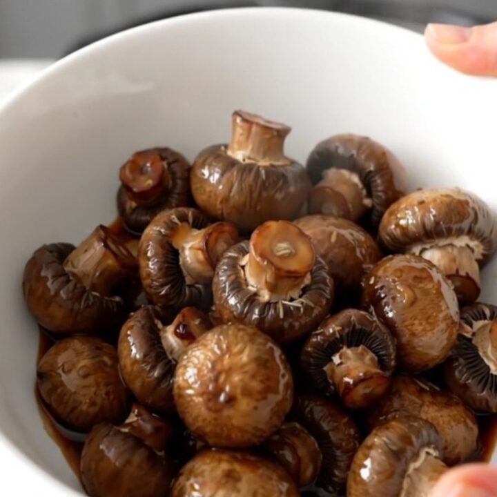 A shallow white bowl filled with dark, wine-braised button mushrooms in a glossy, rich brown buttery sauce.