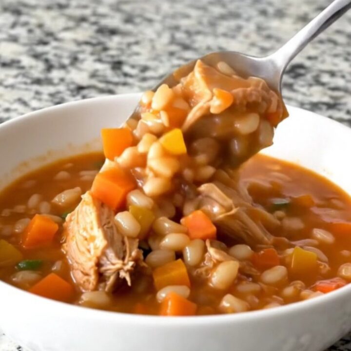 A steaming bowl of turkey and barley stew with visible pieces of carrot, green beans, and corn in a rich tomato broth.