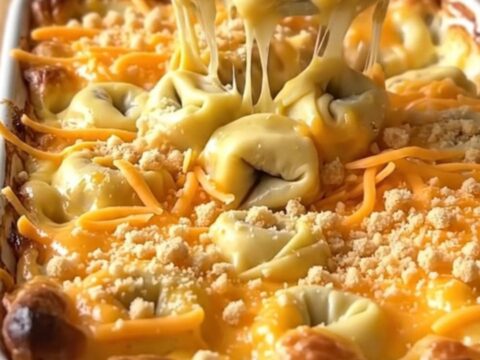 A golden, bubbling tortellini mac and cheese bake in a white ceramic dish on a light wood counter.