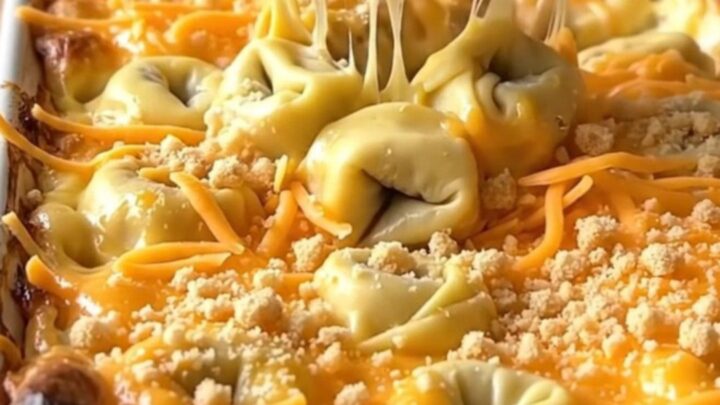 A golden, bubbling tortellini mac and cheese bake in a white ceramic dish on a light wood counter.