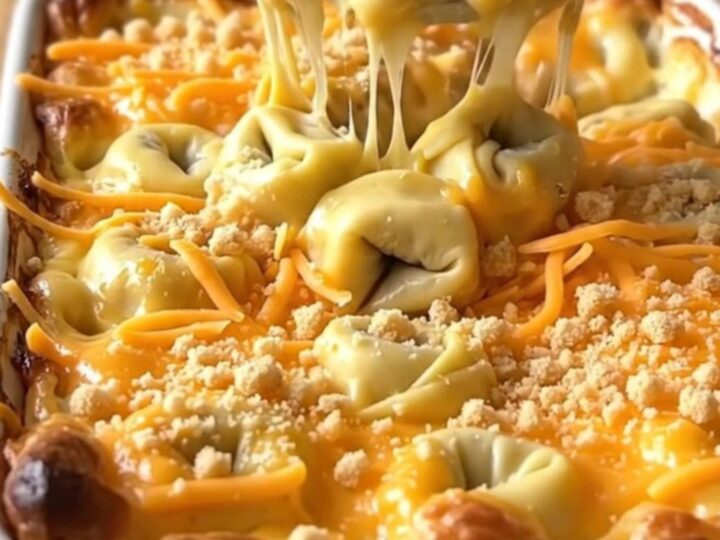 A golden, bubbling tortellini mac and cheese bake in a white ceramic dish on a light wood counter.