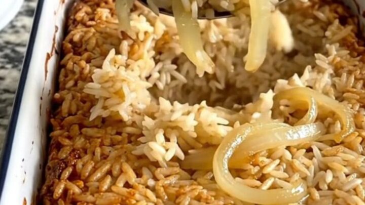 A steaming scoop of golden French onion rice bake lifted from a white rectangular dish.