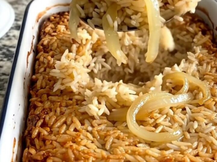 A steaming scoop of golden French onion rice bake lifted from a white rectangular dish.
