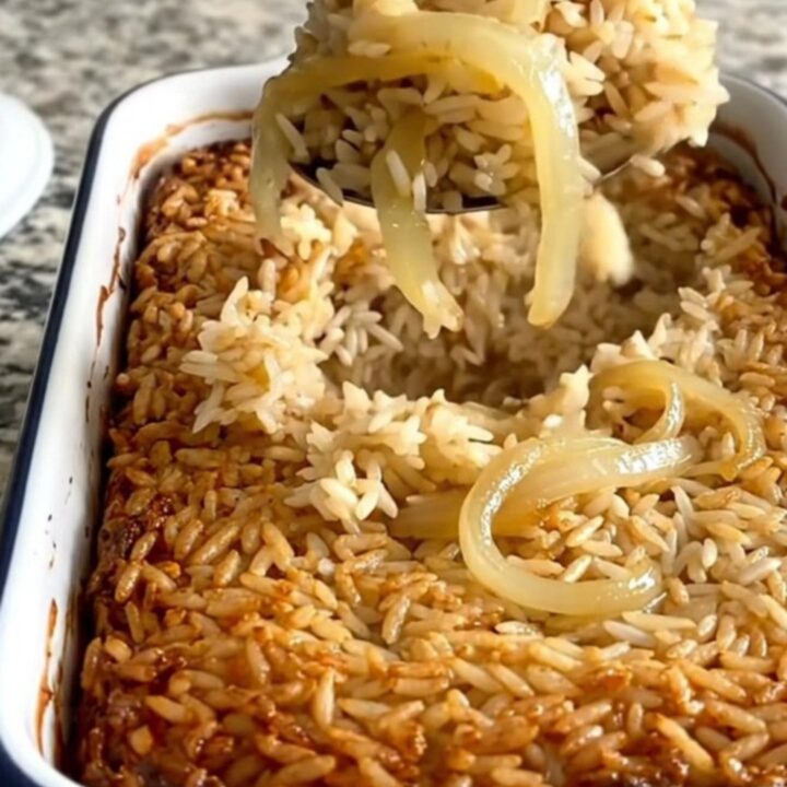 A steaming scoop of golden French onion rice bake lifted from a white rectangular dish.