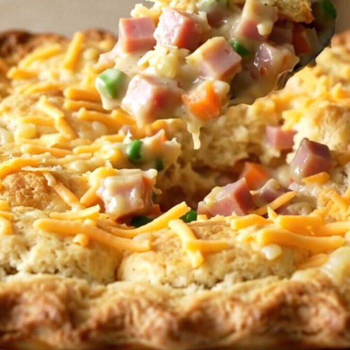 A top-down view of a golden, bubbling ham and cheddar biscuit bake in a white ceramic dish.