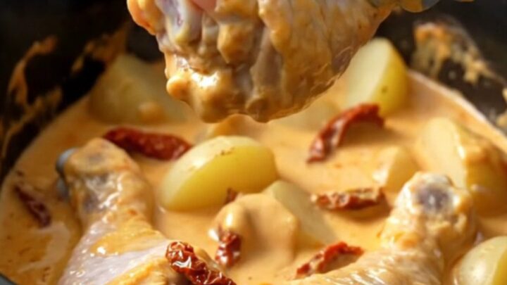 A top-down view of a white ceramic slow cooker filled with chicken drumsticks, halved potatoes, and a creamy sauce.