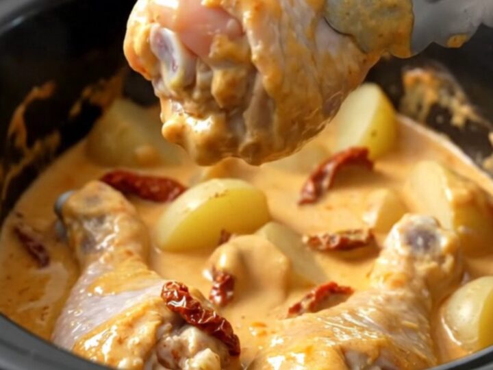 A top-down view of a white ceramic slow cooker filled with chicken drumsticks, halved potatoes, and a creamy sauce.