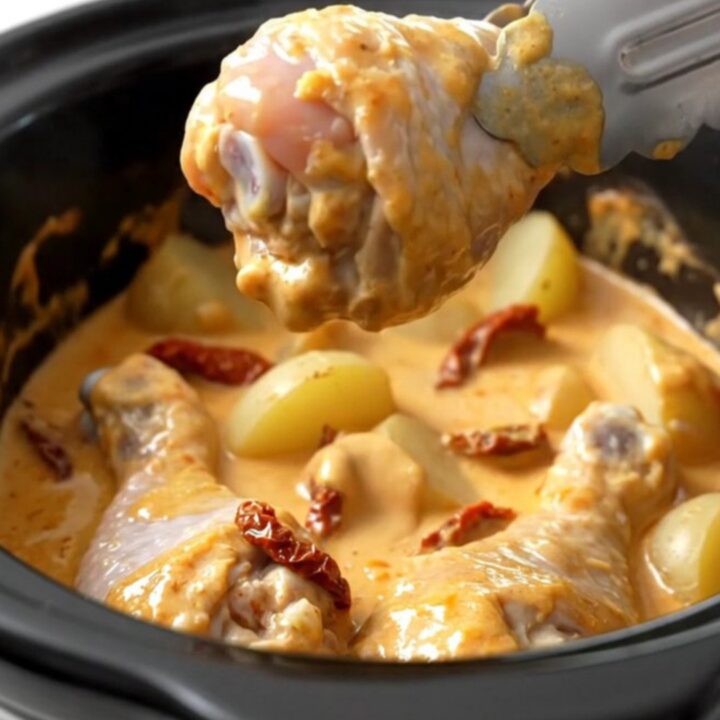 A top-down view of a white ceramic slow cooker filled with chicken drumsticks, halved potatoes, and a creamy sauce.