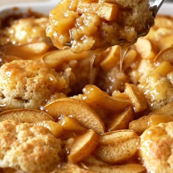 A golden brown apple fritter biscuit bake in a white ceramic dish, with visible apple slices and syrup.