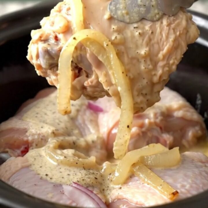 A close-up of tender, braised chicken thighs coated in creamy white barbecue sauce with soft onion slices.