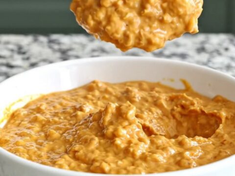 A close-up of creamy barbecue chicken dip in a slow cooker insert, topped with shredded cheddar and barbecue sauce.