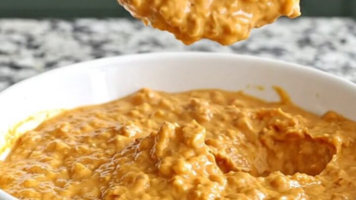 A close-up of creamy barbecue chicken dip in a slow cooker insert, topped with shredded cheddar and barbecue sauce.