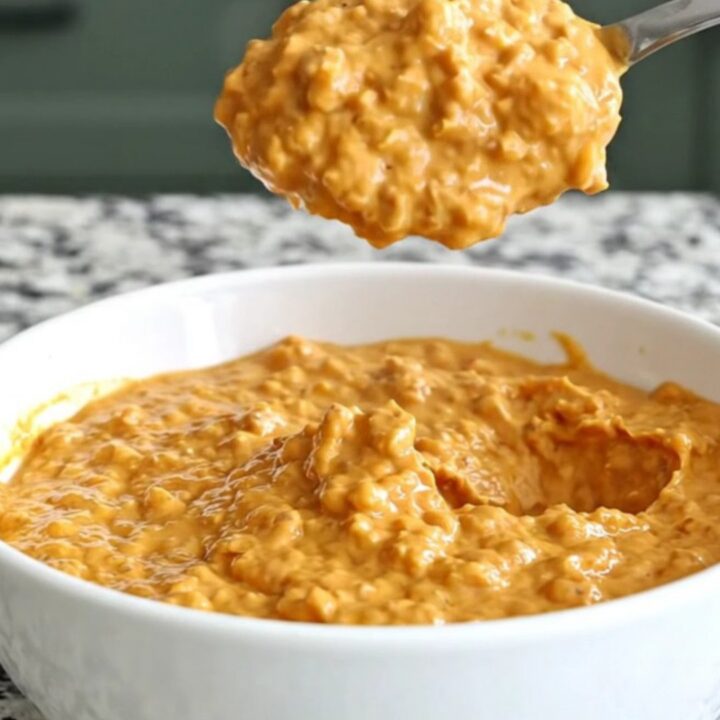 A close-up of creamy barbecue chicken dip in a slow cooker insert, topped with shredded cheddar and barbecue sauce.