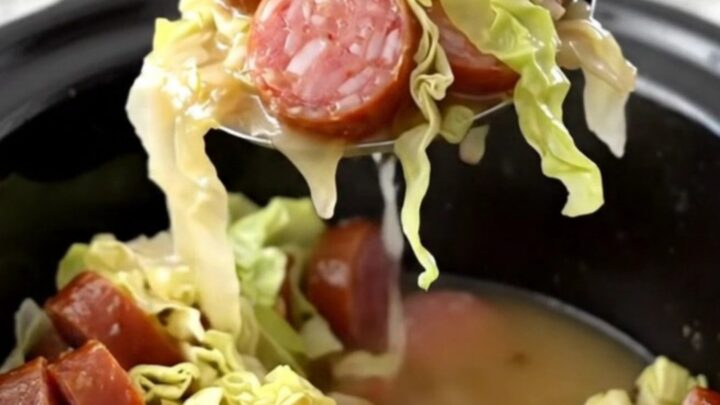 A top-down view of a slow cooker filled with tender green cabbage, coin-shaped smoked sausage, and crispy bacon pieces.