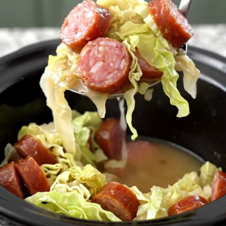 A top-down view of a slow cooker filled with tender green cabbage, coin-shaped smoked sausage, and crispy bacon pieces.