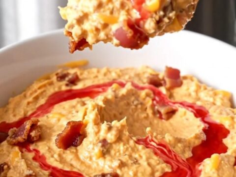 A slow cooker filled with creamy melted cheddar cheese dip topped with bacon crumbles and red pepper jelly swirls.