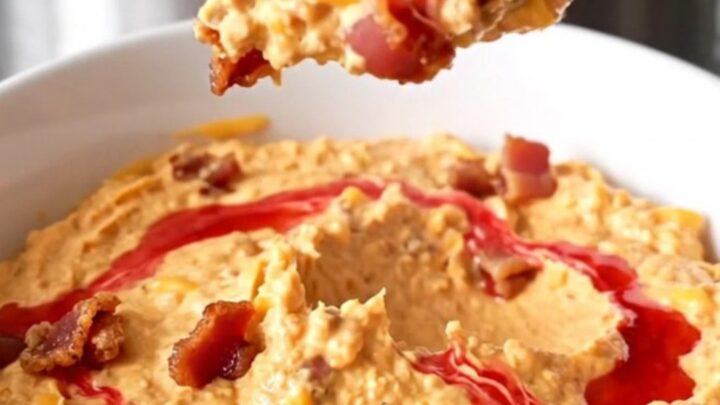 A slow cooker filled with creamy melted cheddar cheese dip topped with bacon crumbles and red pepper jelly swirls.
