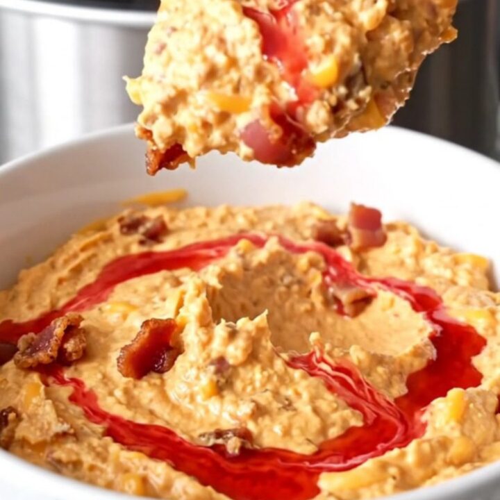 A slow cooker filled with creamy melted cheddar cheese dip topped with bacon crumbles and red pepper jelly swirls.